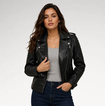 A woman modeling a black faux-leather moto jacket with silver zippers and ribbed sleeves, worn over a gray top and dark blue jeans against a light gray studio background.