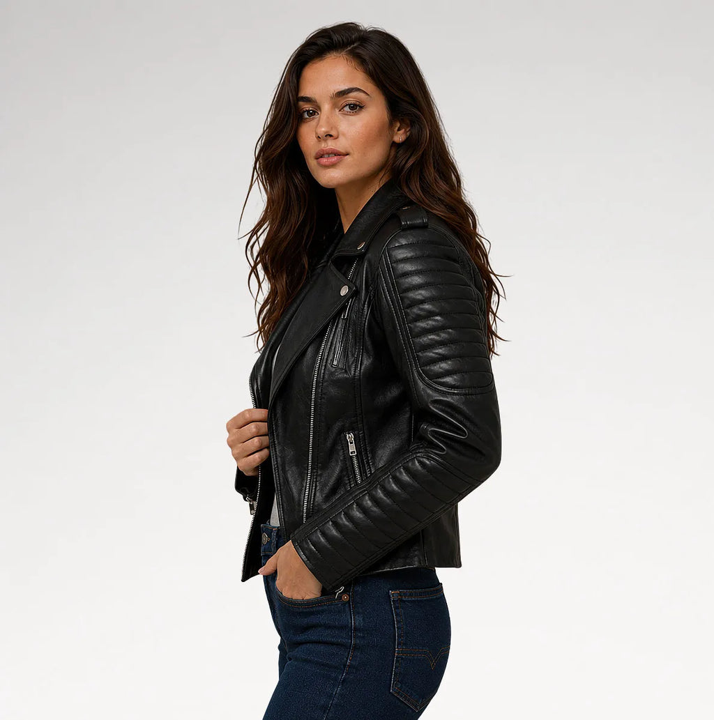 A woman wearing a black leather moto jacket with quilted sleeves and silver zippers, paired with dark blue jeans, photographed against a light gray studio background.