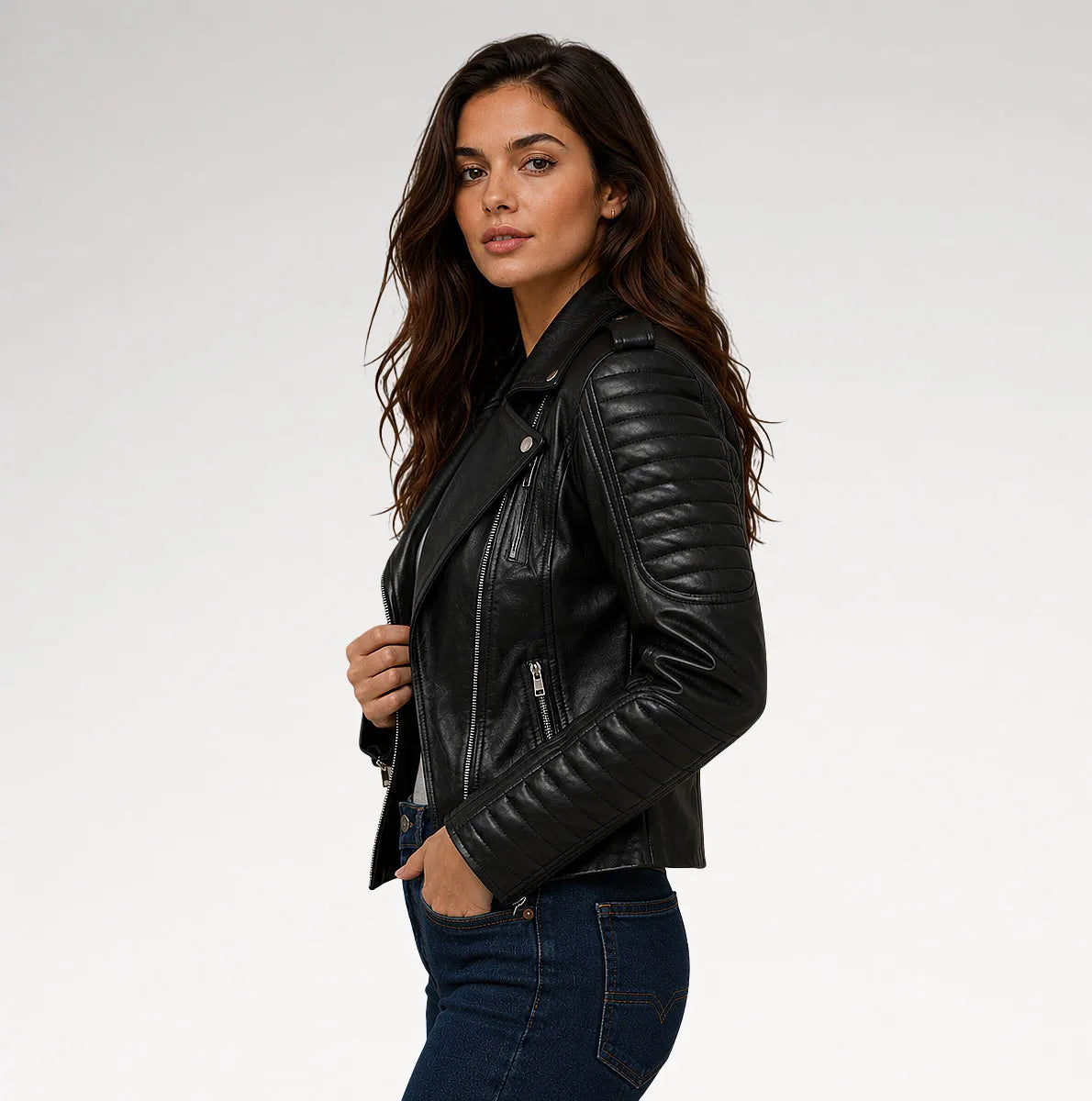 A woman wearing a black leather moto jacket with quilted sleeves and silver zippers, paired with dark blue jeans, photographed against a light gray studio background.