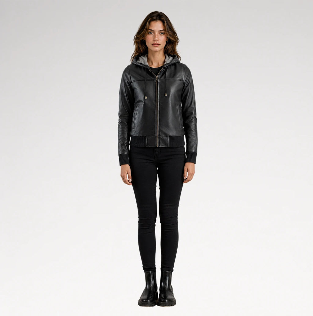 A woman modeling a black faux-leather hooded moto jacket with a front zipper, paired with black skinny pants and black boots, against a light gray studio background.