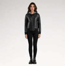 A woman modeling a black faux-leather hooded moto jacket with a front zipper, paired with black skinny pants and black boots, against a light gray studio background.