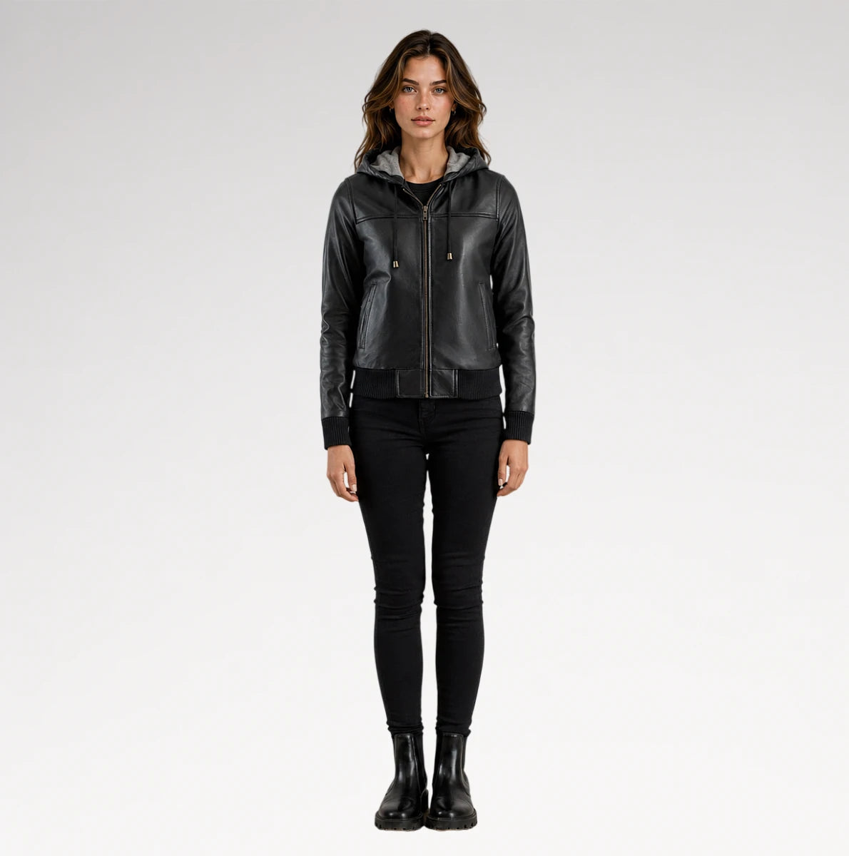 A woman modeling a black faux-leather hooded moto jacket with a front zipper, paired with black skinny pants and black boots, against a light gray studio background.