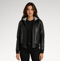 A woman modeling a black faux-leather hooded jacket with a gray fleece-lined hood, worn over a black top and black pants against a light gray studio background.