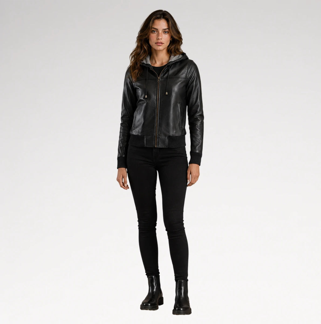 A woman modeling a black leather hooded jacket with gold-tone hardware, paired with black skinny pants and black ankle boots, against a light gray studio background.