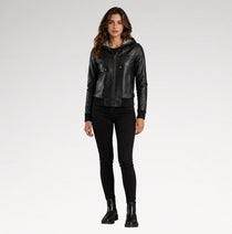 A woman modeling a black leather hooded jacket with gold-tone hardware, paired with black skinny pants and black ankle boots, against a light gray studio background.