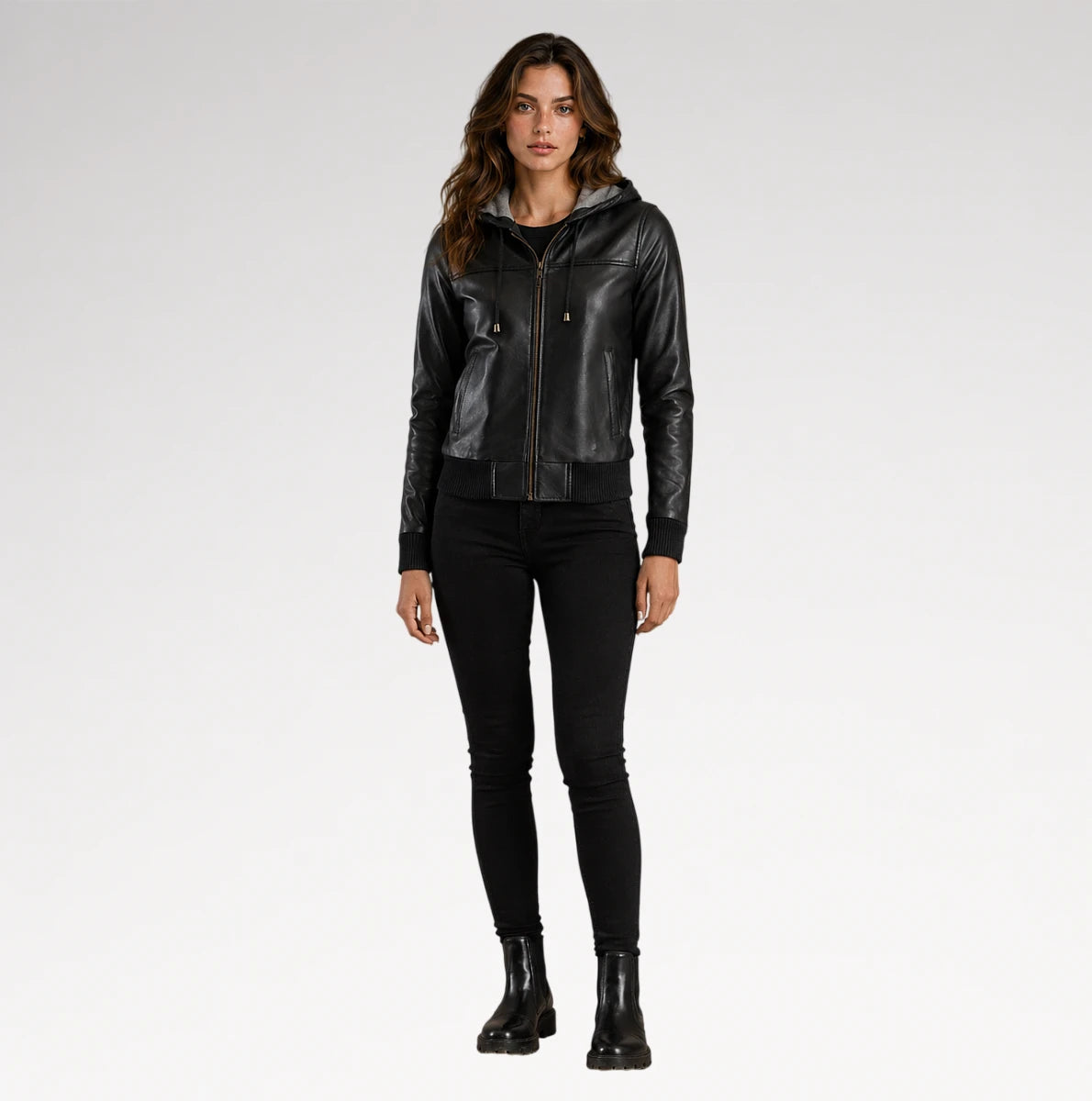A woman modeling a black leather hooded jacket with gold-tone hardware, paired with black skinny pants and black ankle boots, against a light gray studio background.