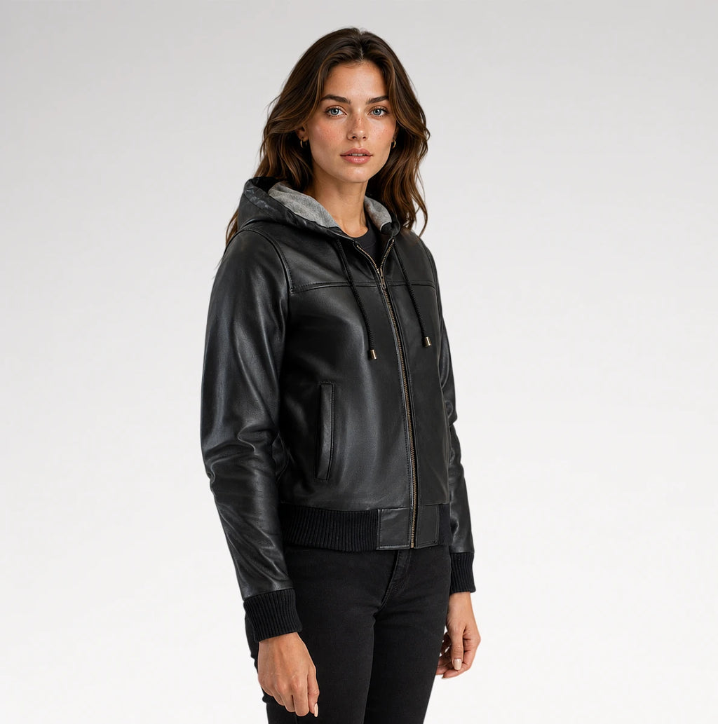 A woman wearing a black faux-leather hooded jacket with a gray hood lining, black ribbed knit trim, and a front zipper, photographed against a light gray studio background.
