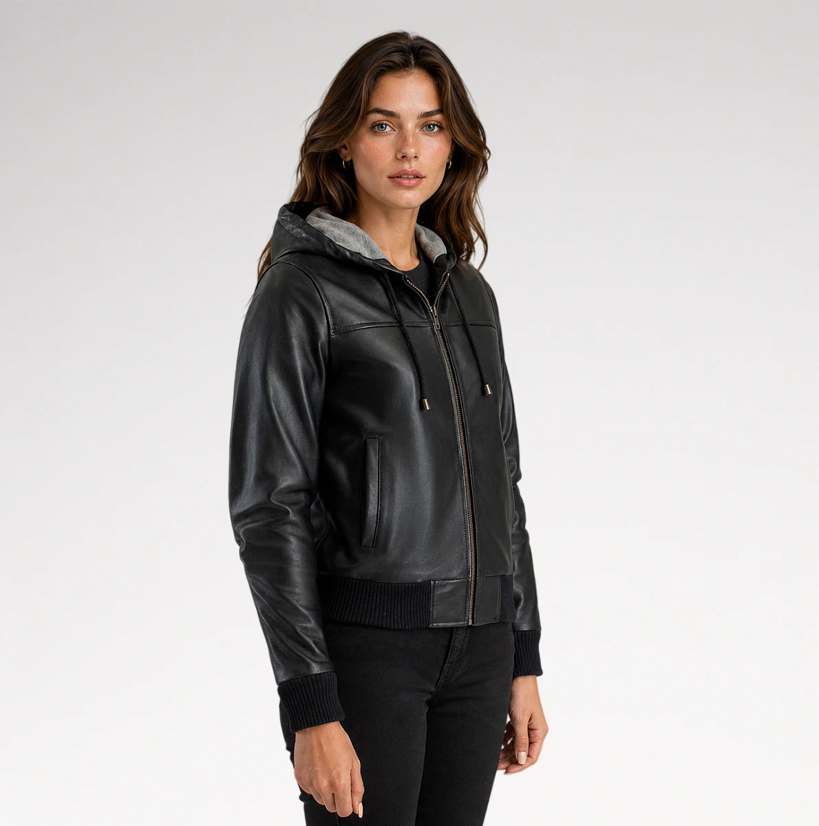 A woman wearing a black faux-leather hooded jacket with a gray hood lining, black ribbed knit trim, and a front zipper, photographed against a light gray studio background.