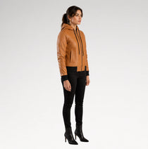 A woman modeling a tan leather hooded jacket with black trim, black pants, and black heeled ankle boots, standing against a light gray studio background.