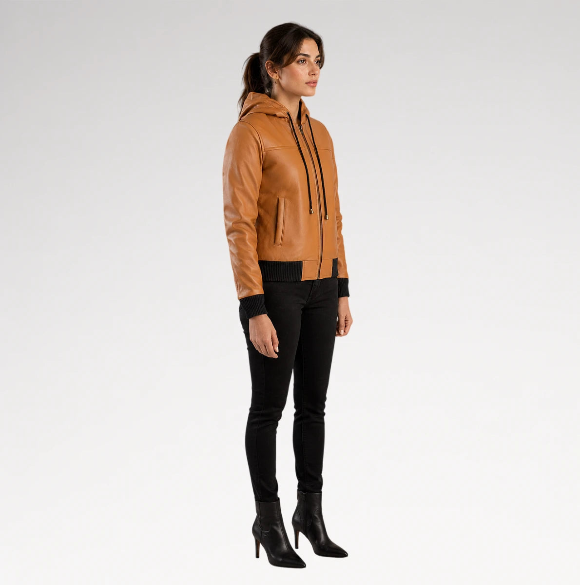A woman modeling a tan leather hooded jacket with black trim, black pants, and black heeled ankle boots, standing against a light gray studio background.