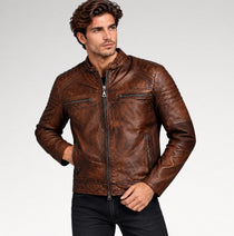 A man wearing a brown distressed leather motorcycle jacket, paired with black jeans, photographed against a light gray studio background.