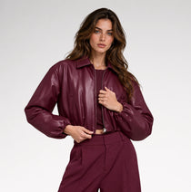 A woman wearing a glossy, deep burgundy faux-leather cropped jacket and matching trousers, photographed from the side against a clean white studio background.