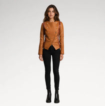A woman modeling a tan leather moto jacket with silver-tone studs and double-breasted buttons, paired with black skinny pants and black ankle boots, against a light gray studio background.