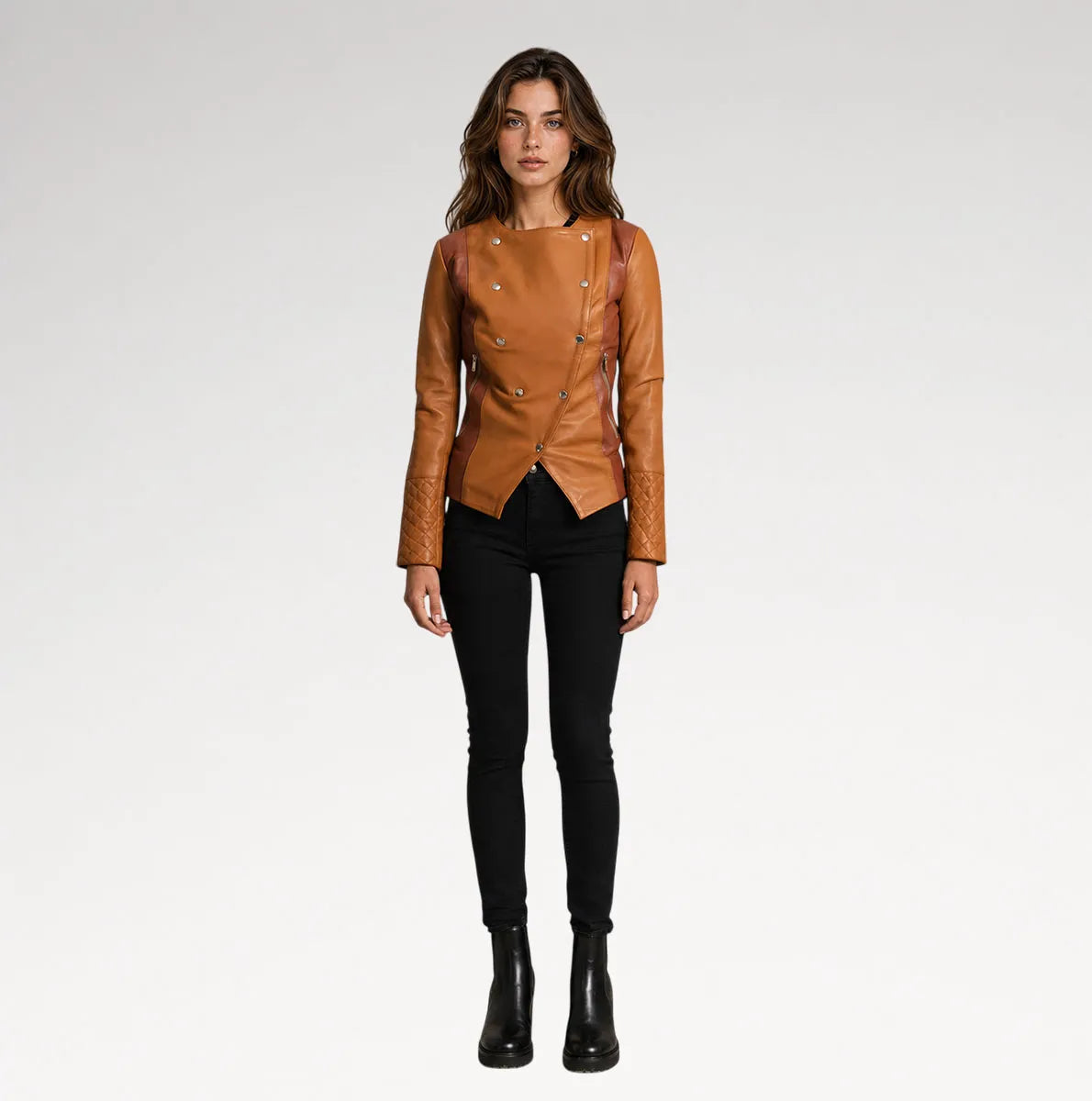 A woman modeling a tan leather moto jacket with silver-tone studs and double-breasted buttons, paired with black skinny pants and black ankle boots, against a light gray studio background.