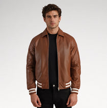 A man wearing a brown leather varsity-style jacket with white stripes on the cuffs, layered over a black t-shirt and black pants, photographed against a light gray studio background.