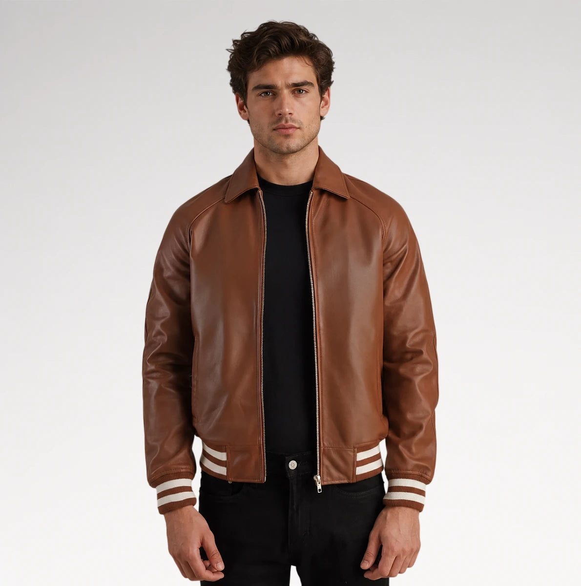 A man wearing a brown leather varsity-style jacket with white stripes on the cuffs, layered over a black t-shirt and black pants, photographed against a light gray studio background.