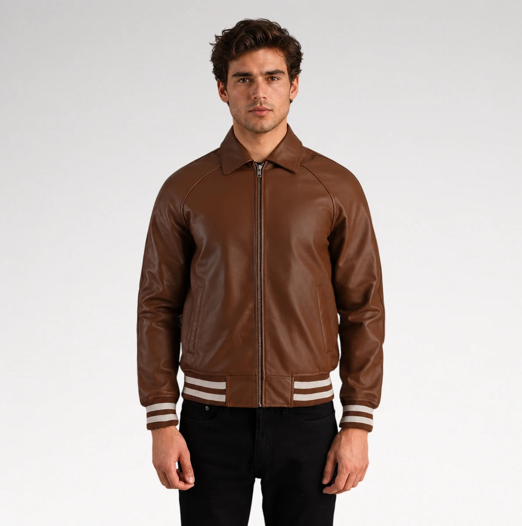 A man wearing a brown leather bomber jacket with cream and black striped ribbed cuffs and hem, paired with black pants, photographed against a light gray studio background.