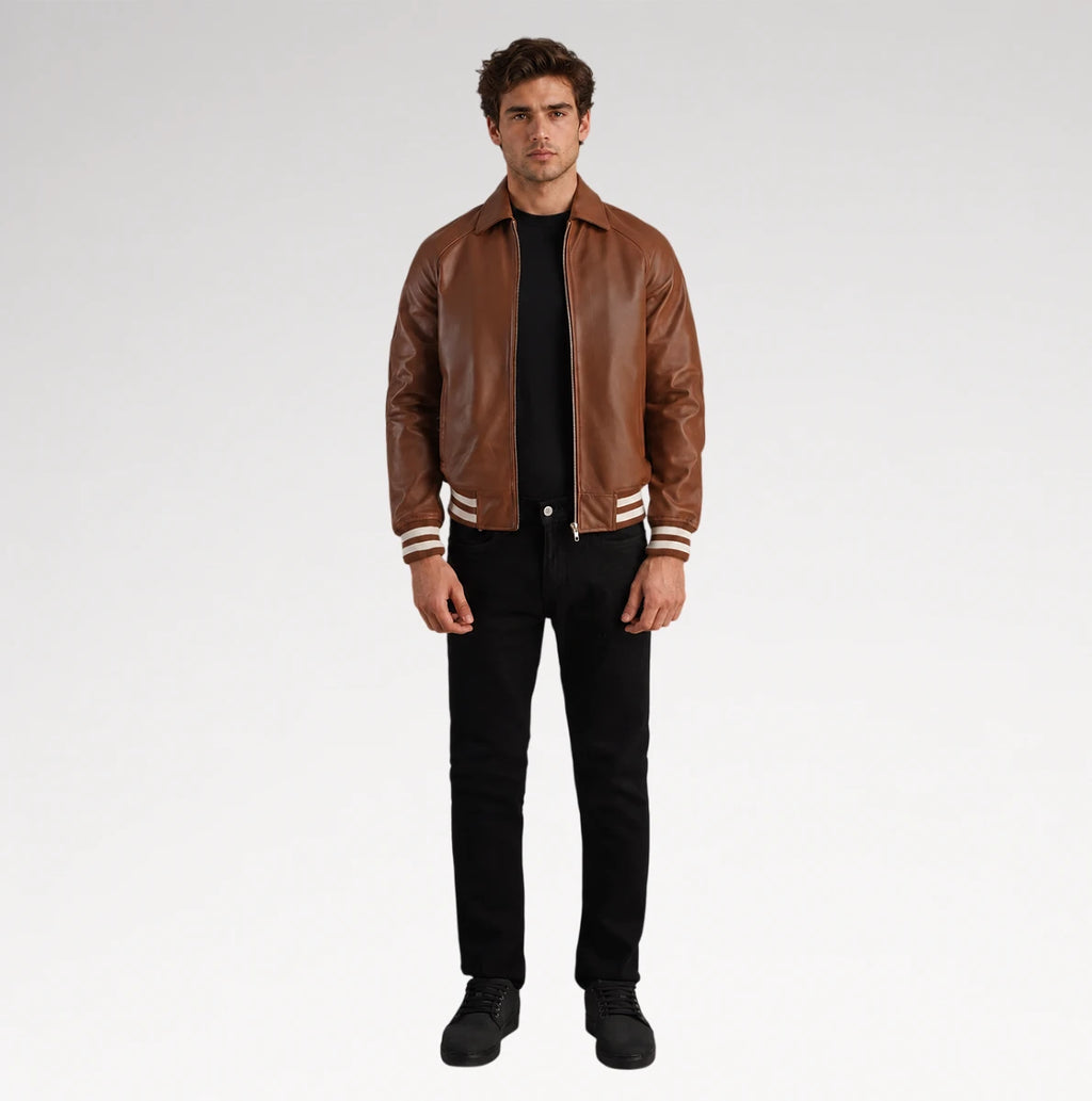 A man modeling a brown leather varsity-style jacket with white stripes on the cuffs and hem, worn over a black shirt and black jeans, against a light gray studio background.