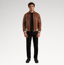 A man modeling a brown leather varsity-style jacket with white stripes on the cuffs and hem, worn over a black shirt and black jeans, against a light gray studio background.
