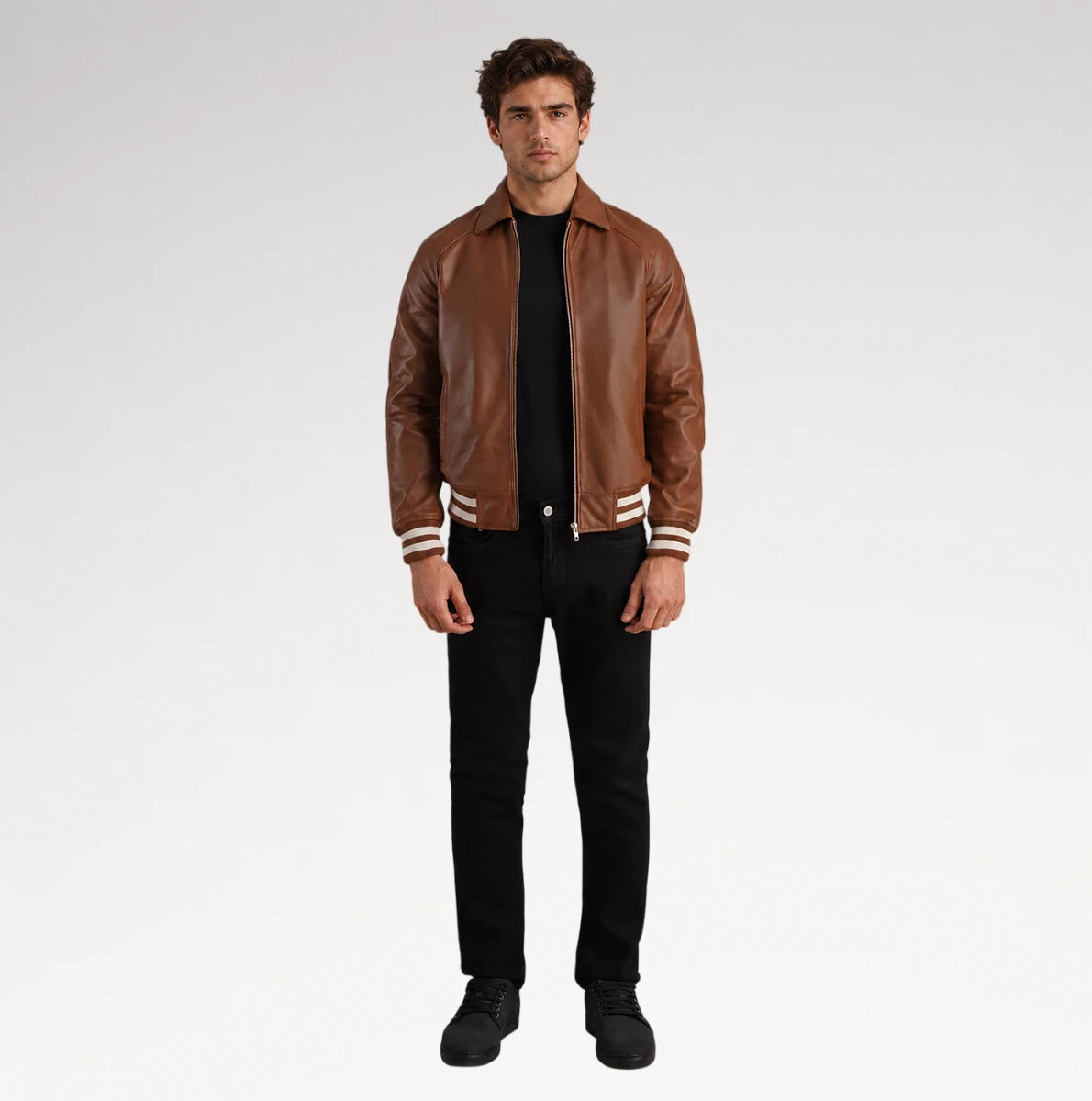 A man modeling a brown leather varsity-style jacket with white stripes on the cuffs and hem, worn over a black shirt and black jeans, against a light gray studio background.