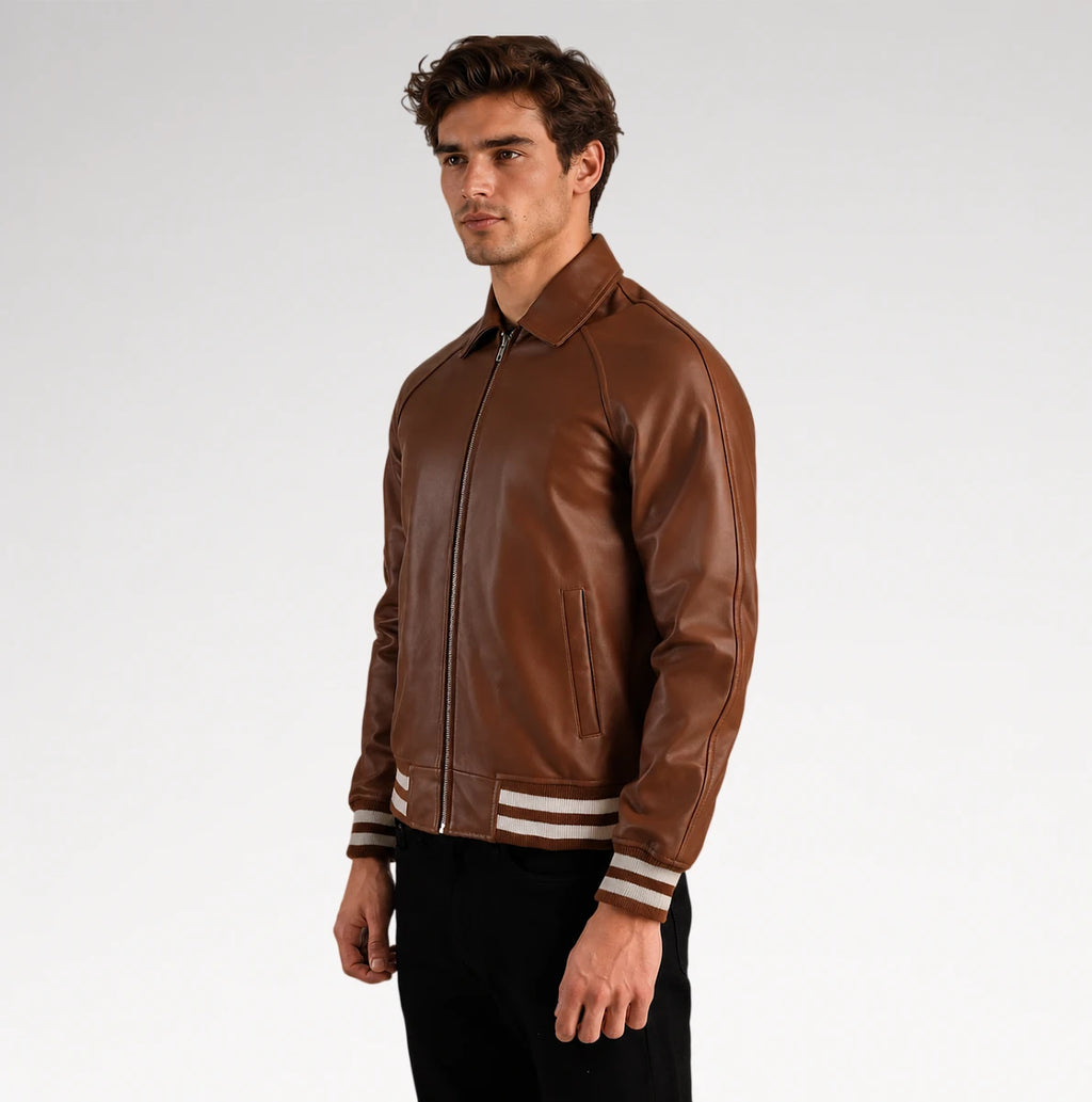 A man wearing a brown leather bomber jacket with white stripes on the cuffs, posed against a light gray studio background.