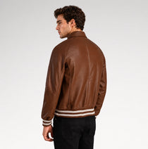 A man wearing a brown leather varsity-style jacket with cream and dark stripes on the ribbed hem and cuffs, shown from a three-quarters back view against a light gray studio background.