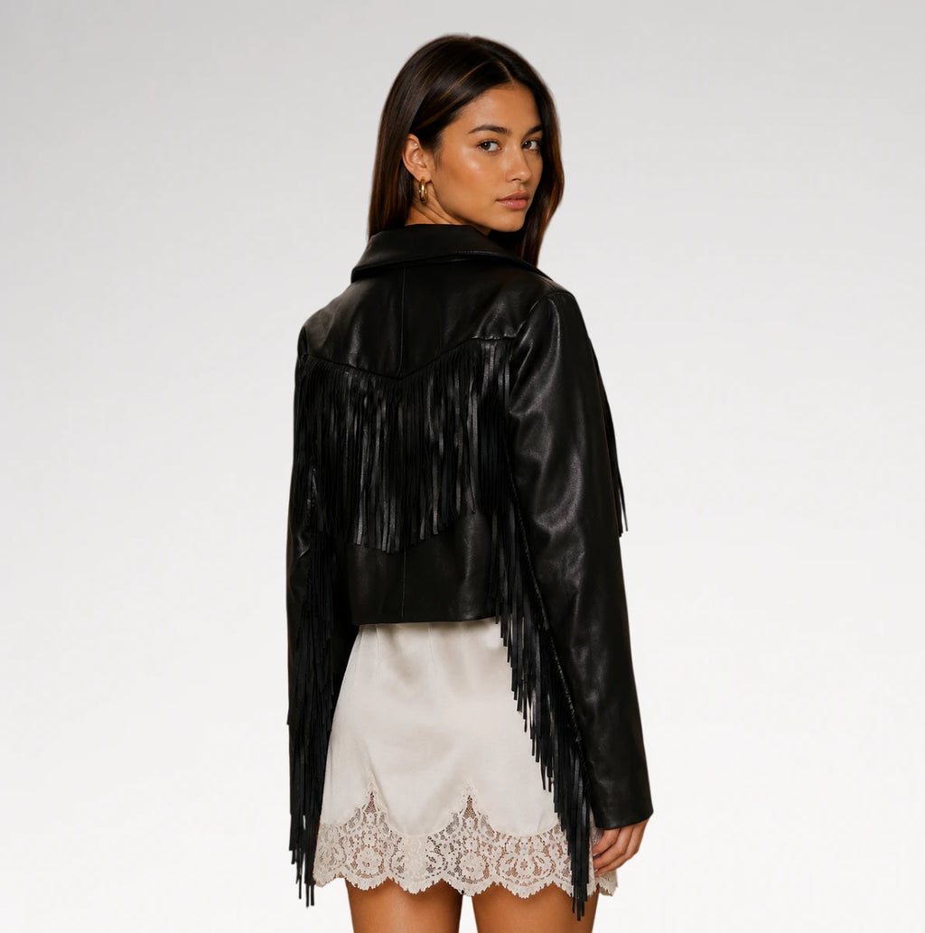 A woman shown from the back wearing a black faux-leather fringe jacket over a white satin skirt with a lace hem, against a plain light gray studio background.
