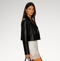 A woman modeling a cropped black faux-leather jacket with a frayed hem, paired with a white lace-trimmed skirt, against a plain light gray studio background.