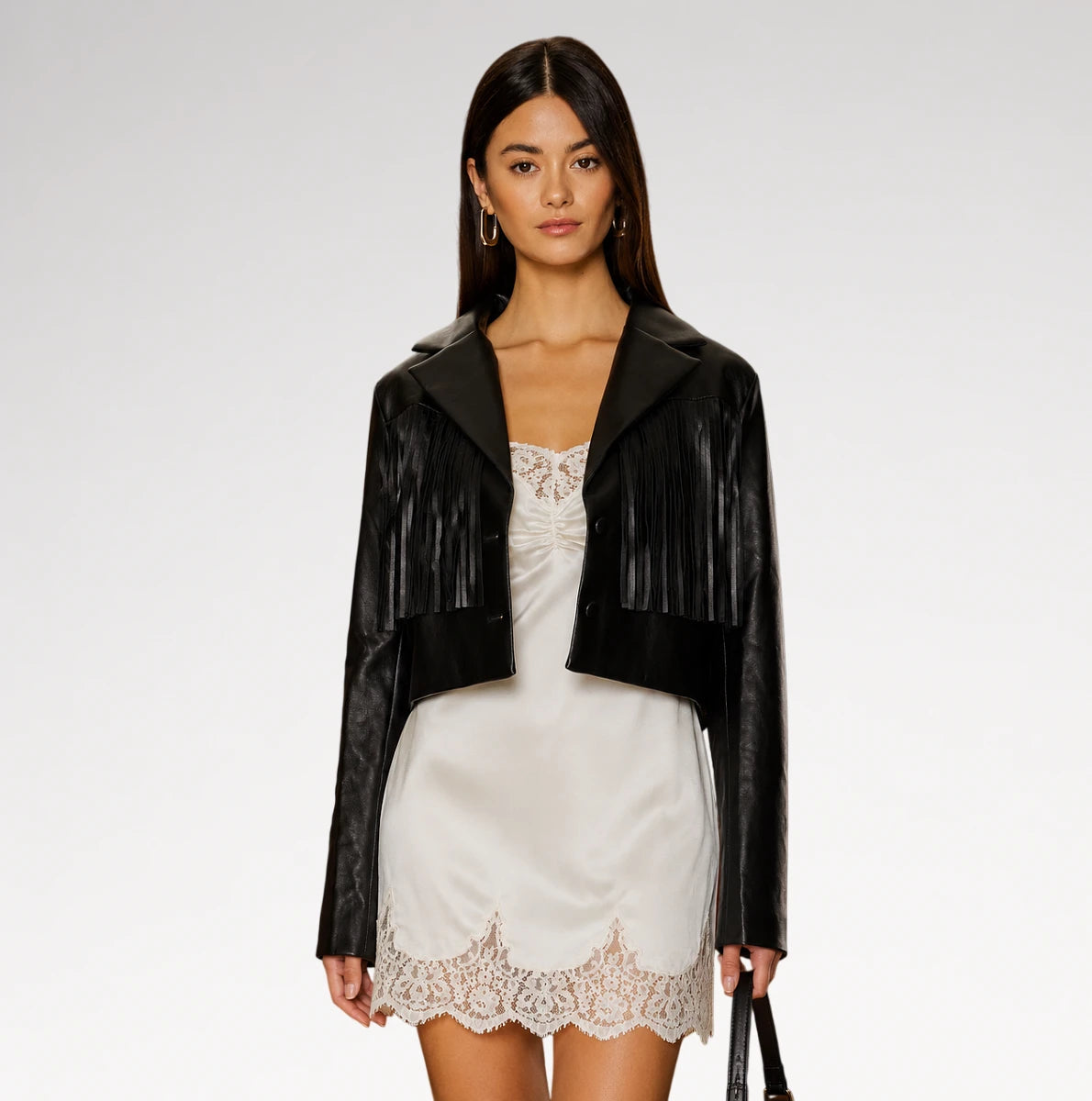 A model wearing a black faux-leather cropped jacket with fringe details over a white satin slip dress, photographed against a light gray studio background. Main image