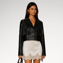 A model wearing a black faux-leather cropped jacket with fringe detailing and a white mini skirt with a lace hem, standing against a light gray studio background.