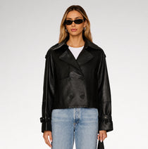 A woman modeling a black leather double-breasted jacket with a white t-shirt and light blue jeans against a light gray studio background.