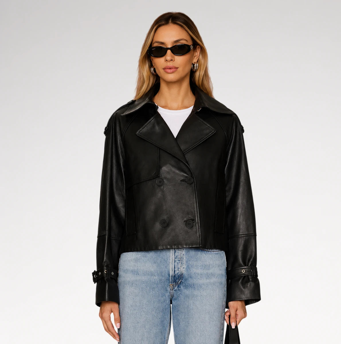A woman modeling a black leather double-breasted jacket with a white t-shirt and light blue jeans against a light gray studio background.