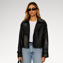 A woman modeling a black leather moto jacket over a white t-shirt and light blue jeans, wearing black sunglasses, against a light gray studio background.