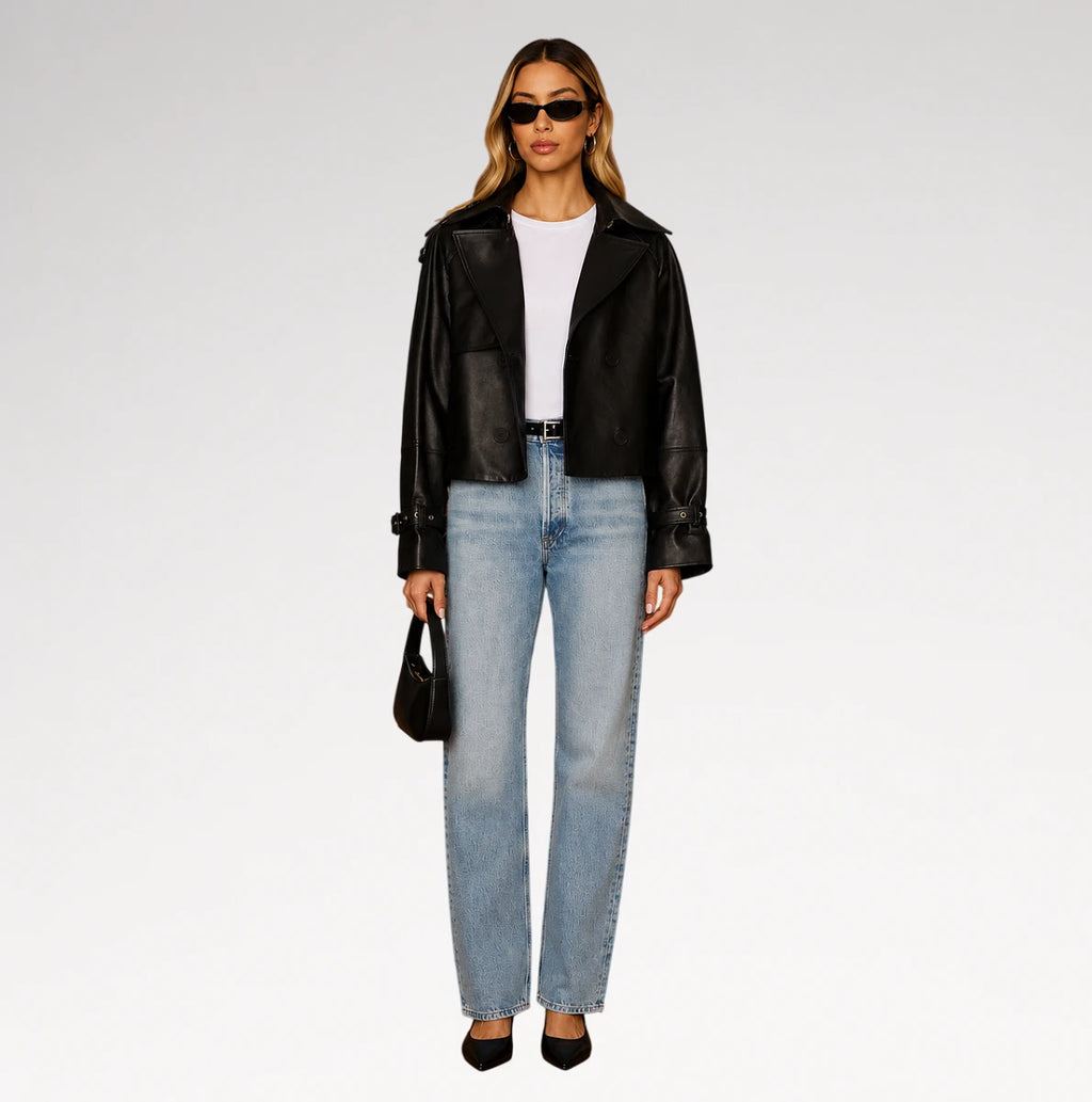 A model stands against a light gray studio background wearing a black leather moto jacket, light-wash blue jeans, a white top, black heels, and holding a small black handbag.