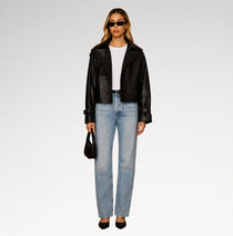 A model stands against a light gray studio background wearing a black leather moto jacket, light-wash blue jeans, a white top, black heels, and holding a small black handbag.
