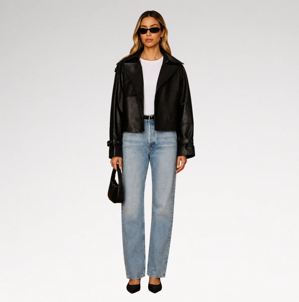 A model stands against a light gray studio background wearing a black leather moto jacket, light-wash blue jeans, a white top, black heels, and holding a small black handbag.