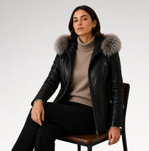 A woman sits on a wooden chair wearing a black leather jacket with a large gray fur-trimmed hood over a beige ribbed turtleneck sweater and black pants, photographed against a light gray studio background.