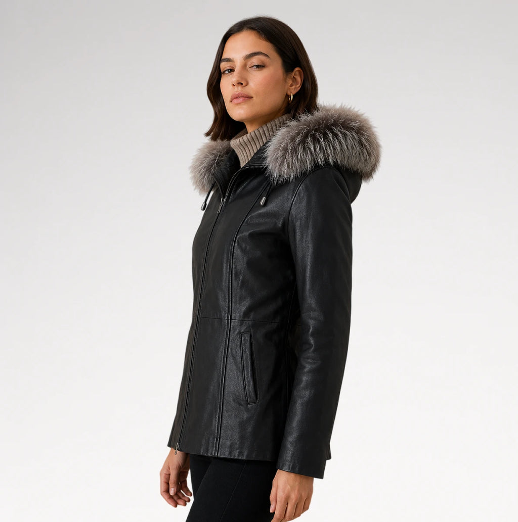 A woman wearing a black faux-leather parka jacket with a large gray fur-trimmed hood, photographed against a plain light gray studio background.