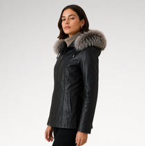 A woman wearing a black faux-leather parka jacket with a large gray fur-trimmed hood, photographed against a plain light gray studio background.