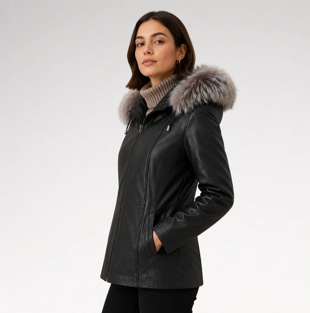 A woman wearing a black leather jacket with a large gray fur-trimmed hood, standing against a light gray studio background.