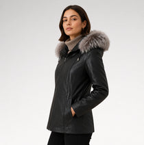 A woman wearing a black leather jacket with a large gray fur-trimmed hood, standing against a light gray studio background.