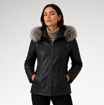 A woman wearing a black faux-leather parka jacket with a large gray faux-fur trimmed hood, styled over a beige ribbed turtleneck sweater and black pants, photographed against a light gray studio background.