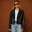 Woman wearing a black leather jacket, white top, and blue jeans against a brown background