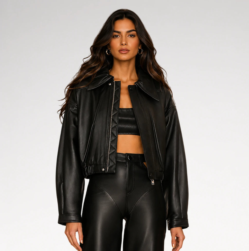 A woman modeling a black faux leather moto jacket, matching black crop top, and black faux leather pants against a light gray studio background.