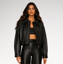A woman modeling a black faux leather moto jacket, matching black crop top, and black faux leather pants against a light gray studio background.