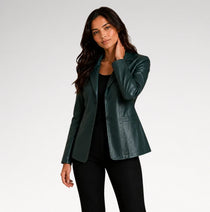 A woman modeling a dark green faux leather blazer over a black top and black pants, photographed against a light gray studio background.