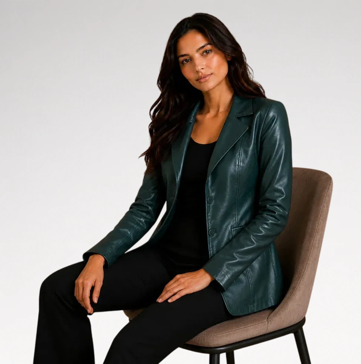 A woman sits on a brown upholstered chair wearing a dark teal-green faux leather blazer over a black top and black pants, photographed against a light gray studio background.