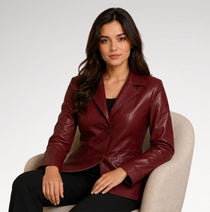 A woman seated in a light beige armchair wearing a fitted leather blazer in deep burgundy with a black top and black pants, photographed against a clean light gray studio background.