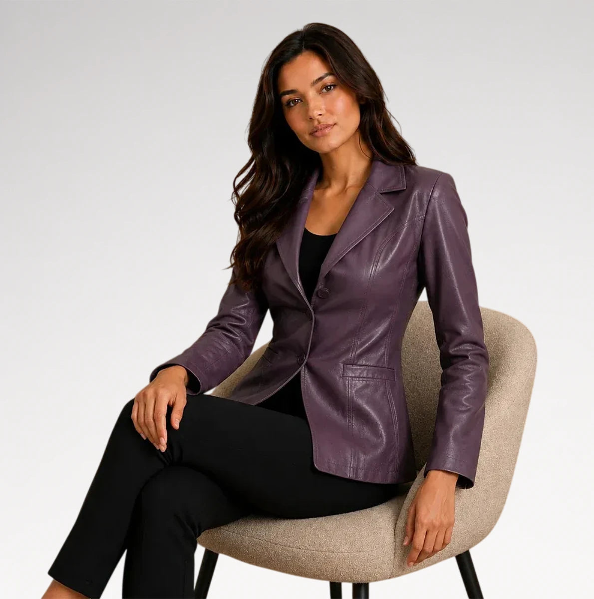 A woman sits on a beige upholstered chair wearing a fitted purple faux-leather blazer over a black top and black pants, photographed against a light gray studio background.
