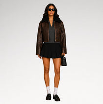 A model stands against a light gray studio background wearing a brown leather jacket with a black shearling-style collar, a dark gray knit top, a black pleated mini skirt, white socks, black loafers, sunglasses, and carrying a small black handbag.